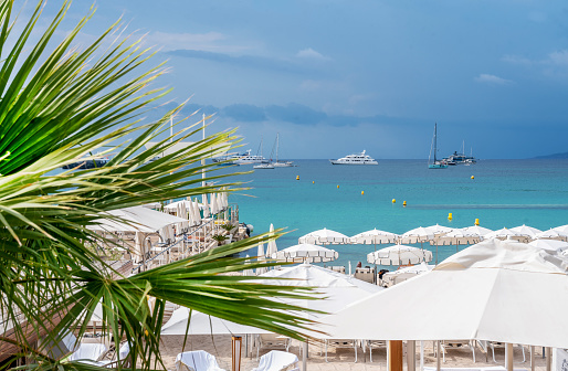 One of the private beaches on the Croisette with white beds (chaise lounges) and tents and yachts anchored in the bay (save money) in Cannes, Gorgeous view of the Acoustre Riviera with palm leaves