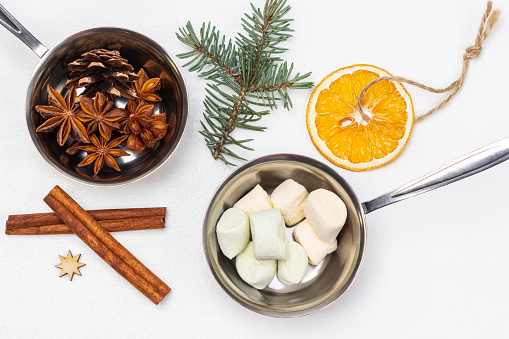 Marshmallow and star anise in metal bowls. Cinnamon sticks, a sprig of spruce and a dry orange on a string. Flat lay. White background.
