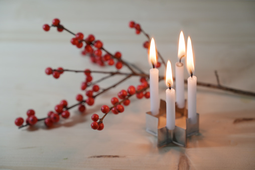 Mini advent wreath, four small candles on a cookie cutter in star shape in front of a holly branch with red berries on a wooden table, selected focus, narrow depth of field