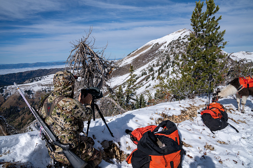 Wyoming, USA - October 2, 2022: Hunter wearing camo uses a spotting scope to view deer during a hunt in the snow