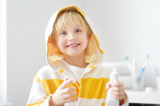 Cute blonde caucasian child applying cream on his face in bathroom in the morning. Kids care cosmetics. Hygiene for little children