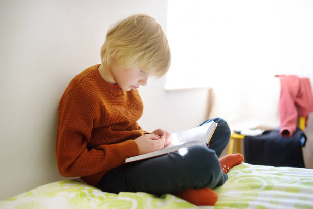 Smart preteen boy sitting on bed and enthusiastically reading interesting book. Kid reader enjoying interesting stories, reading fascinating adventurous novels. Child loving to read. Bookworm