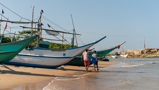 Galle, Sri Lanka - November 15, 2019: three fishing boats are parked on the shore of the Atlantic Ocean, near the fish market in Hikkaduwa. Local fishermen are walking around the boat.