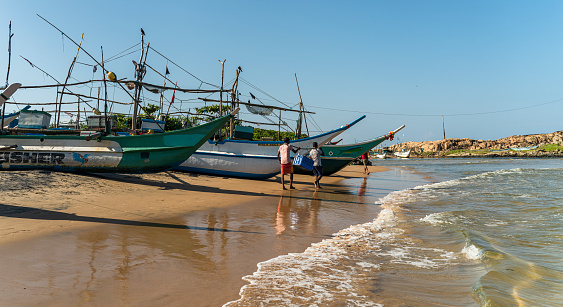 Galle, Sri Lanka - November 15, 2019: three fishing boats are parked on the shore of the Atlantic Ocean, near the fish market in Hikkaduwa. Local fishermen are walking around the boat.