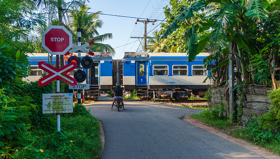 Galle, Sri Lanka - November 15, 2019: two boys on a bicycle are waiting on the road for a passenger train to pass. There is a stop sign in front of the traffic lights.