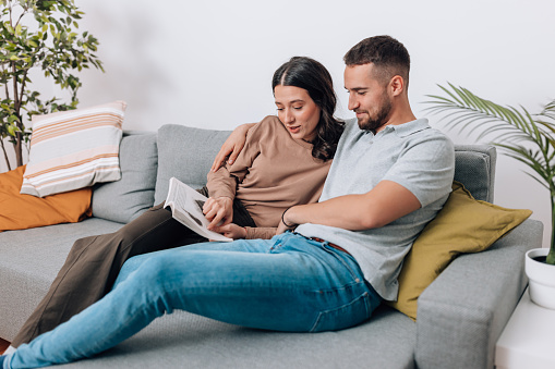 A young Caucasian couple is sitting on a sofa and reading a book with smiles on their faces.