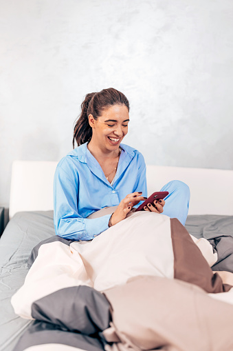 A young Caucasian woman wearing pajamas is sitting on her bed and laughing while checking her cellphone.