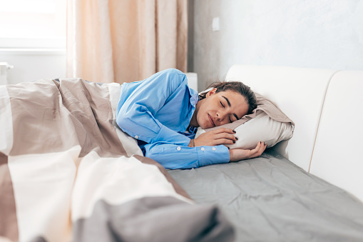 A young Caucasian woman is lying under her bed covers with the morning sun coming through the window.