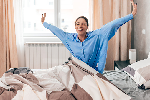 A young Caucasian woman wearing pajamas is cheerfully stretching in her bed with a smile on her face.