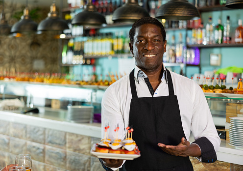 Positive African American waiter serving tapas for friends meeting for drinks and food in the pub