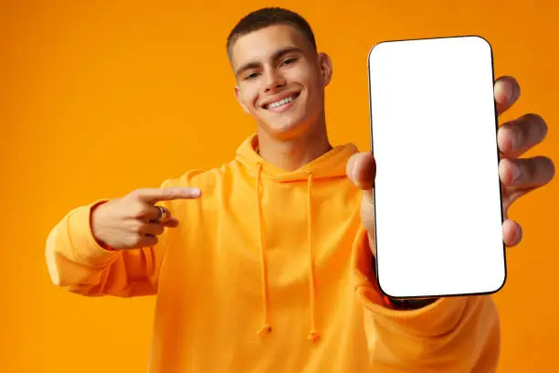 Smiling young man showing mobile phone blank white screen on yellow background copy space