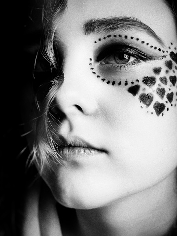 Beautiful teen girls face, half shadowed with a serious expression and wearing creative make up with hearts and designs around the eyes. Black and white close up portrait.