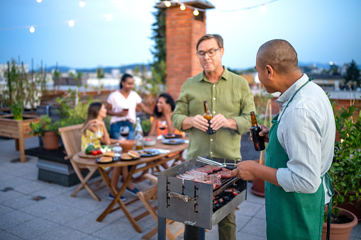 Men having a beer and grilling a meat steak while women having conversation at the rooftop dinner party