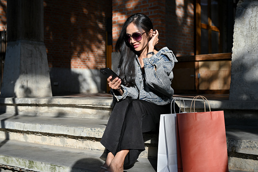 Gorgeous young Asian woman in sunglasses with her shopping bags using her smartphone while sitting on street stairs. Urban city lifestyle and shopping concept