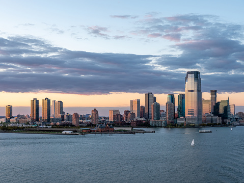 Idyllic landscape showing Jersey City and Hudson River (USA). The image is illuminated by the setting sun. A beautiful cloudy sky with warm summer colours. In the foreground, on the Hudson River, we can see boats and sailboats. The bottom of the sky is fire-coloured.