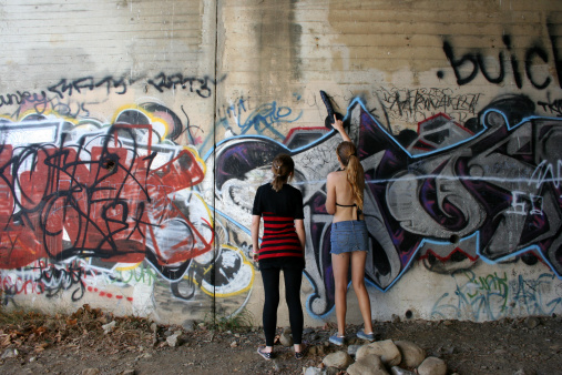 two teenaged girls stand in front of a grafitti covered wall.