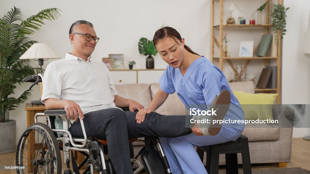 asian female care attendant helping senior man on wheelchair doing rehab training. she slowly lifts his injured leg while he shows painful face in the living room at home Home Caregiver Stock Photo