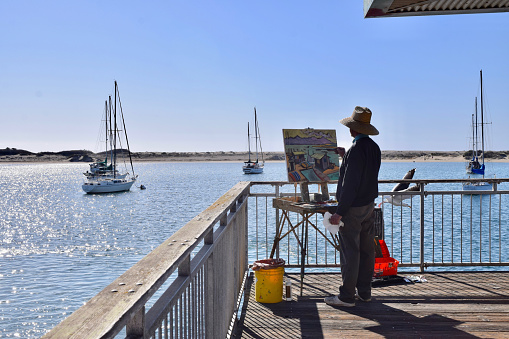 Fresno, United States – March 04, 2021: A photo of a Fine Art Artist painting outside on his canvas on a Pier out into the water and boats against a blue clear sunny sky