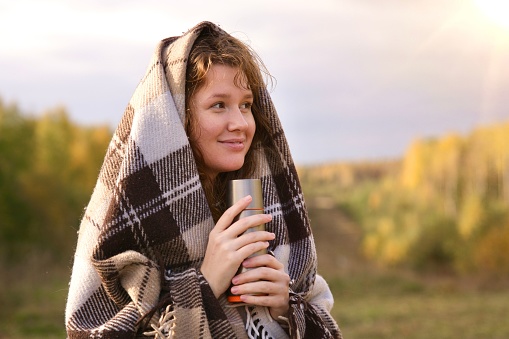 A portrait of a young smiling Caucasian woman covering herself with a blanket and holding a thermo bottle on autumn trees background