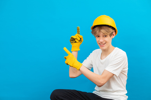A teenage boy in a yellow helmet is looking at the camera and points fingers up, standing in a studio on a blue background. The concept of construction and repair