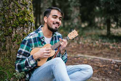 A bearded Hispanic man wearing flannel singing while playing ukulele under a tree