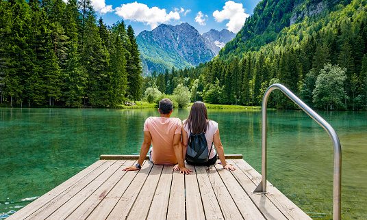 Rear view of young female sitting on wooden deck by beautiful lake in mountains. Green, spring, flower crown, outdoors. Zgornje Jezersko, Slovenia