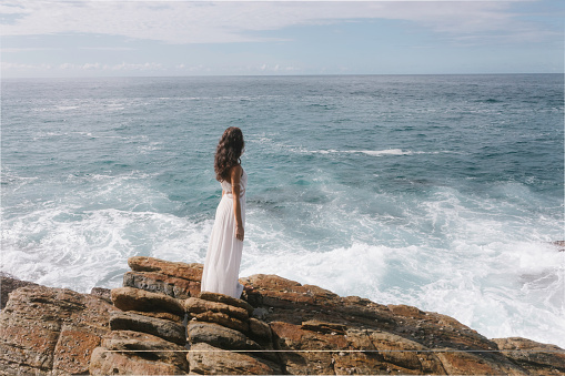 Aerial drone view of beautiful bride in an amazing white dress standing on a cliff and looking at the sea. Epic scene with stormy waves and gorgeous woman model