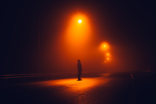 Moody dark image depicting a strange sinister man back lit by street lamps in an abandoned parking lot.