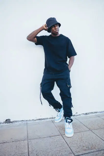 A vertical shot of a young black man in street-style clothes posing leaning on a white wall outdoors