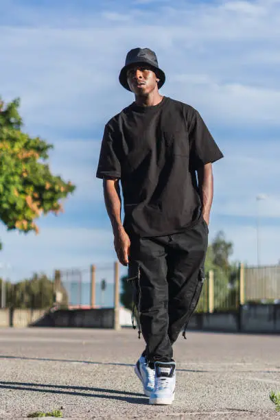 A vertical shot of a young black man in street-style clothes walking outdoors on a sunny day