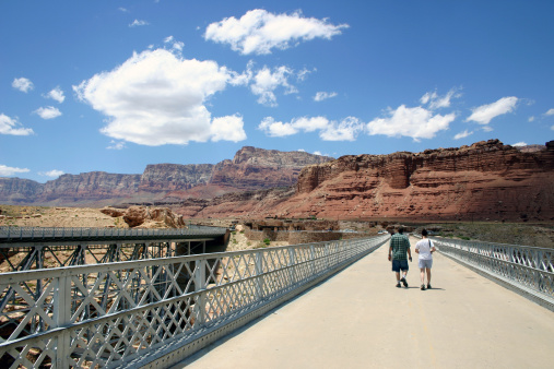 Young couple hike the Navajo Bridge over the Colorado River at Marble Canyon. The Vermillion Cliffs before them and the Navajo Nation behind. Arizona, USA. 