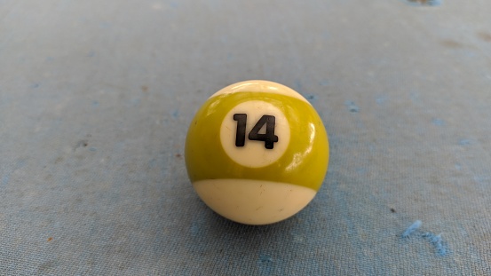 The billiard ball showing the number 14. The ball is on the pool table. There are defects around the ball due to play.
