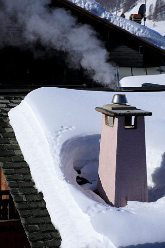 Snowy Rooftop Smoking Winter Chimney.  White smoke comes out of a house's chimney on a winter day.  \nHigh mountain   Italian Alps  ski area. Ski resort Livigno. italy, Europe.
