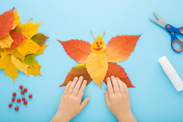 istock Child hands creating butterfly shape from colorful leaves on light blue table background. Pastel color. Toddler development. Making autumn decorations. Point of view shot. Closeup. Top down view. 1442139153
