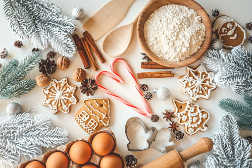High angle view of a white table filled with ingredients for preparing and baking gingerbread man cookies. The composition includes a baking sheet, a rolling pin and ingredients like eggs, flour, cinnamon, brown sugar, cloves, butter among others.