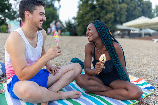 A portrait of a beautiful young multiracial couple eating ice cream and sitting on the seaside of the river bank.