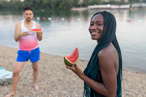 A young African woman and her male friend are enjoying watermelon on a warm sunny day near the water.