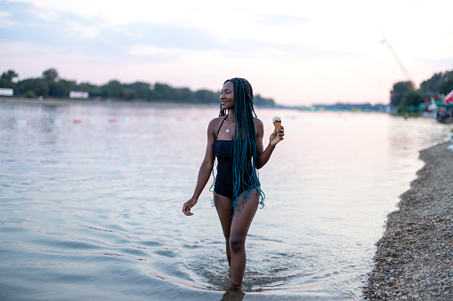 A young woman of African ethnicity in a swimsuit enjoys a summer day walking in the river and refreshing with cold ice cream.