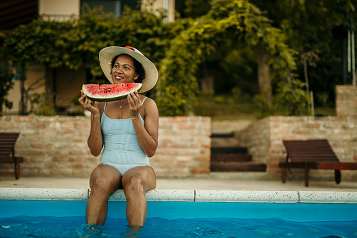 Shot of a beautiful young woman relaxing with legs in a swimming pool and eating fruits.