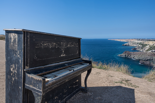 An old vintage piano on top of a cliff at Cape Fiolent in Crimea. Close-up against the blue sky. A picturesque mountain range that adorns the coastline of the sea. Water of various shades of blue.