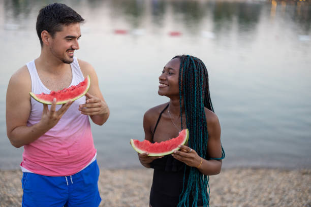 A young multiracial couple near a river is eating watermelons. A young African woman and her male friend are enjoying watermelon on a warm sunny day near the water. black man eating watermelon stock pictures, royalty-free photos & images