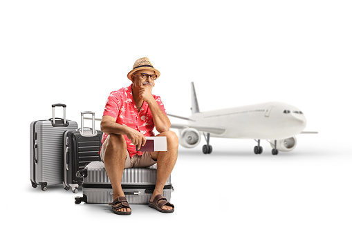 Mature male toruist sitting on a suitcase and waiting in front of an airplane isolated on white background