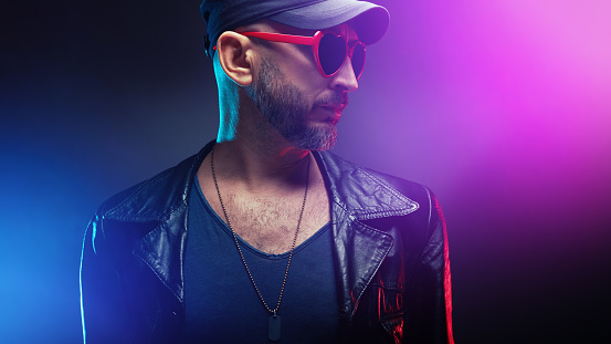 Stylish handsome man wearing leather jacket under neon club lights with heart shaped red sunglasses