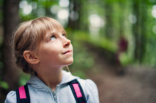Portrait of a little girl hiking with backpack. The girl aged 6 is looking up at the beautiful trees in the forest.\nNikon D700