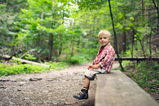 Cute little boy aged 3 hiking in the forest. The boy is resting on a bench and smiling at the camera.
Shot with Nikon D700