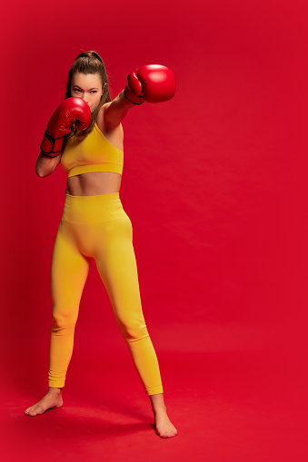 Portrait of young woman in yellow sportswear, boxing, training posing isolated over red background. Developing strength. Concept of youth culture, emotions, facial expression, sport, lifestyle, health