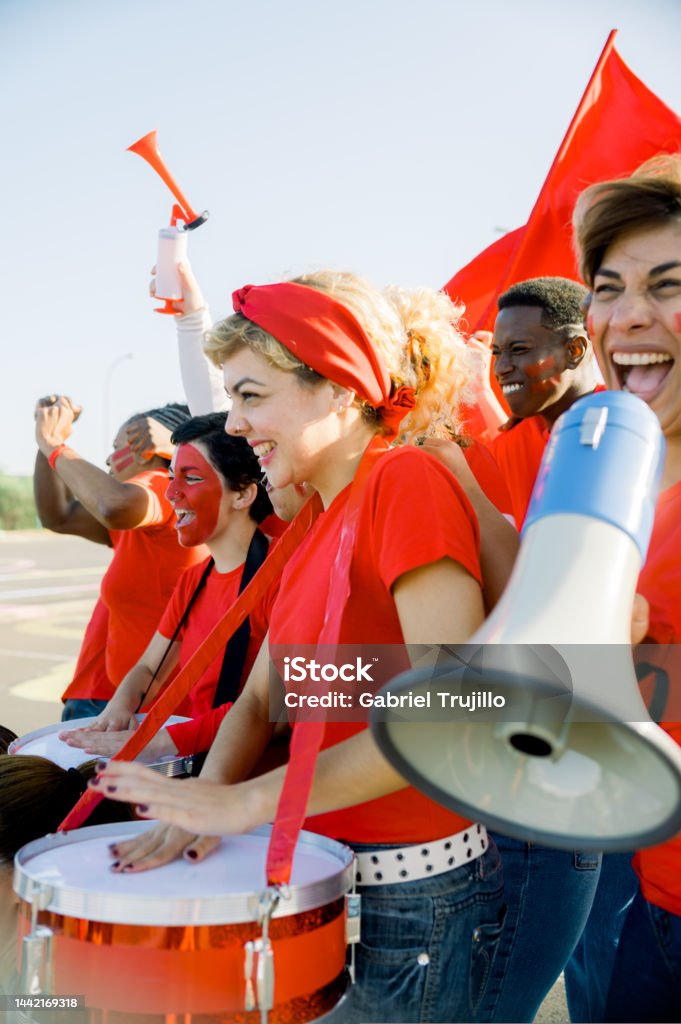 Excited football fans beating drums and yelling while walking on street Delighted young multiracial male and female football fans in red t shirts and painted faces smiling and shouting happily while playing drums and celebrating win after soccer match Fan - Enthusiast Stock Photo