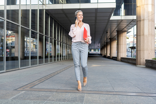 Cheerful business woman holding documents and talking via smart phone when walking outdoors
