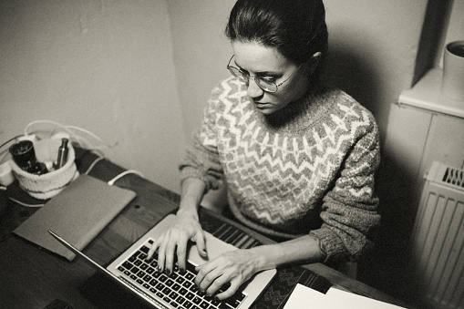 Portrait of a woman at her workplace at home. She is working intently on her laptop until late at night. She is wearing a warm sweater and the heater can be seen behind her.