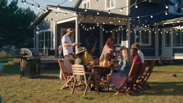 Family and Multiethnic Diverse Friends Gathering Together at a Garden Table. People Cooking Meat on a Fire Grill, Preparing Tasty Salads for a Big Family Celebration with Relatives.